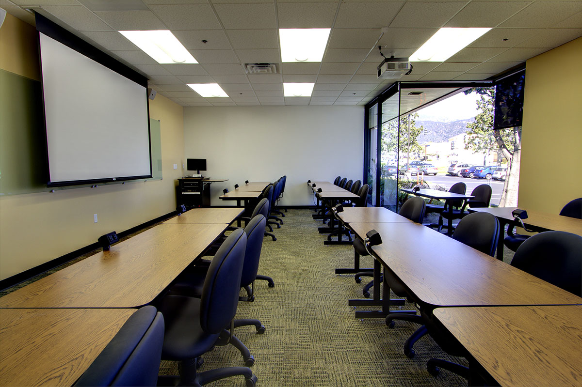 A conference room with black swivel chairs, brown tables, and a big white screen in front.
